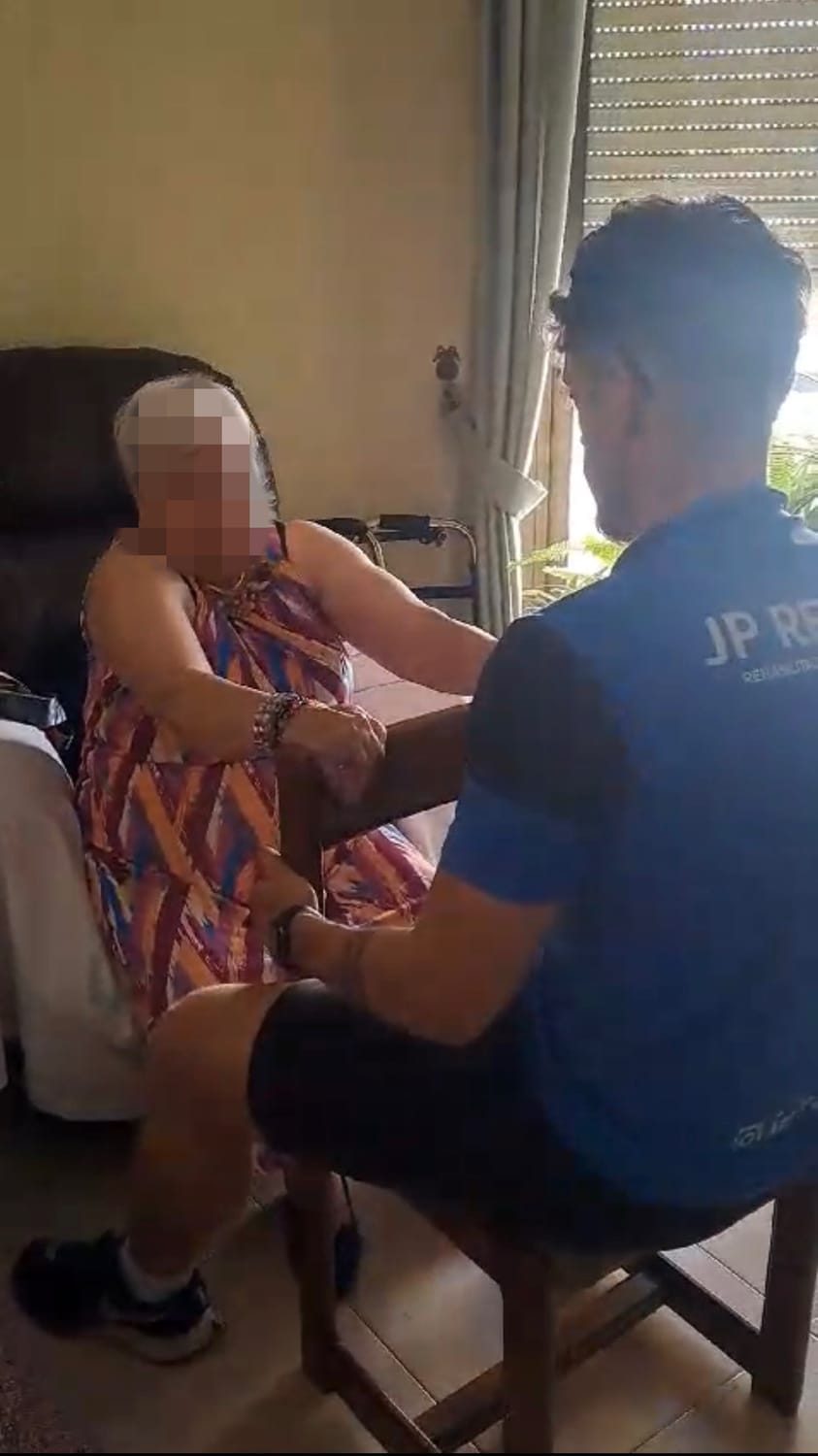 Sesión de fisioterapia con una paciente mayor sentada en su salón